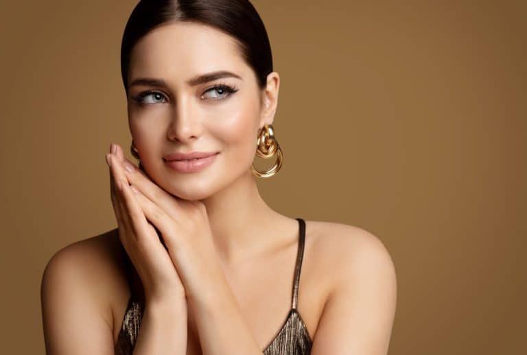 Brunette woman with light blue eyes wearing gold earrings. Her hair is in a bun and her hands are placed gently clasped beneath her neck as she looks out of frame to the right. The background is gold. 