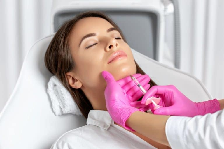 Woman with eyes closed on a medical table with a towel beneath her head. Her eyes are closed and a pair of orange gloved hands coming in from the right of the frame inject a solution into her chin. 
